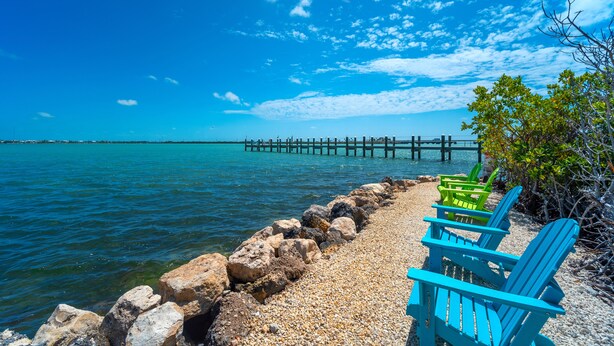 A pebbled beach with blue Adirondack chairs and a wooden pier extending into the water. - Haven House (Little Torch Key)