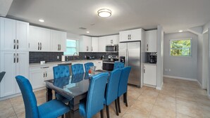 A modern kitchen with white cabinets, a stainless steel refrigerator, and a dining area with blue chairs. - Haven House (Little Torch Key)