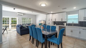 A modern kitchen with white cabinets, a dining area with blue chairs, and a large window with blinds. - Haven House (Little Torch Key)