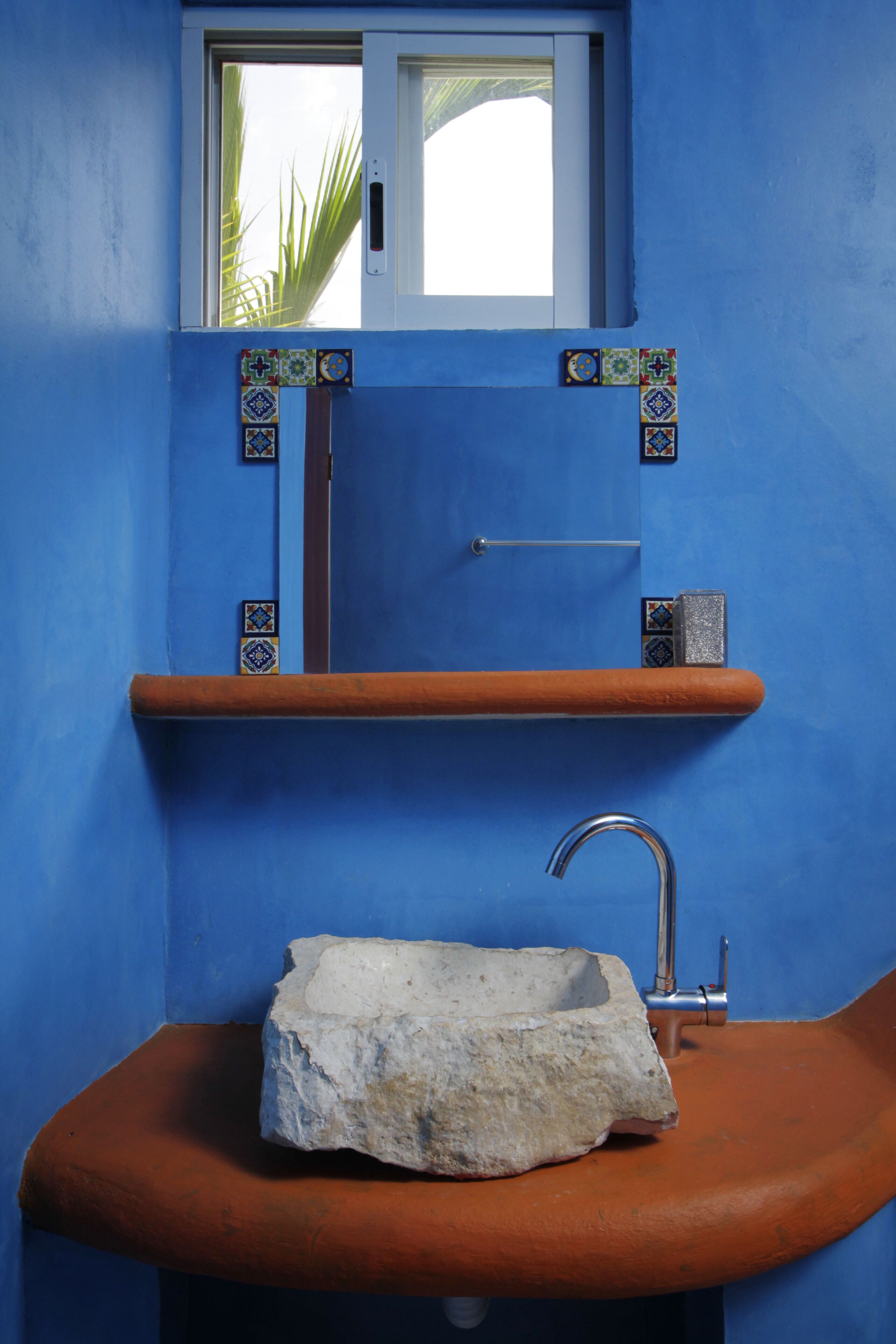 A bathroom with a blue wall, a window, a sink with a stone basin, and a shelf with tiles.