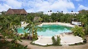 A swimming pool surrounded by tropical vegetation, with a thatched-roof building and a white building with multiple windows in the background.