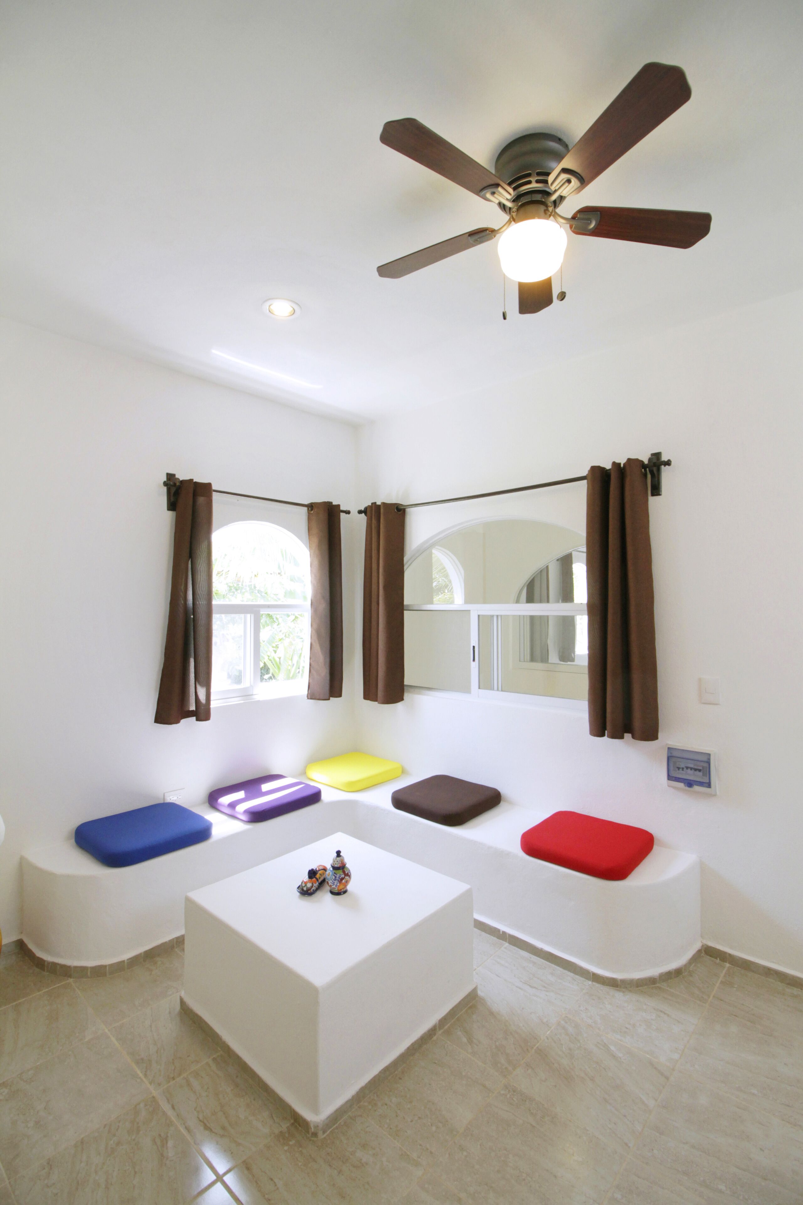 A room with a ceiling fan, a window with curtains, and a seating area with colorful cushions.