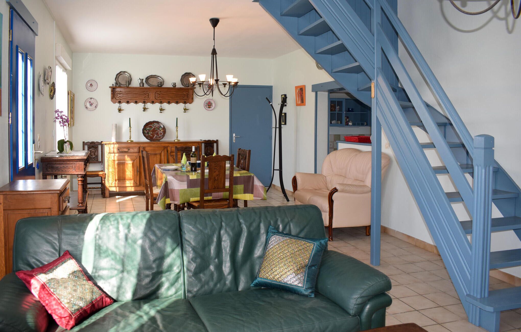 A living room with a green leather sofa, a dining area with a wooden table and chairs, a staircase, and a kitchen area with blue cabinets.