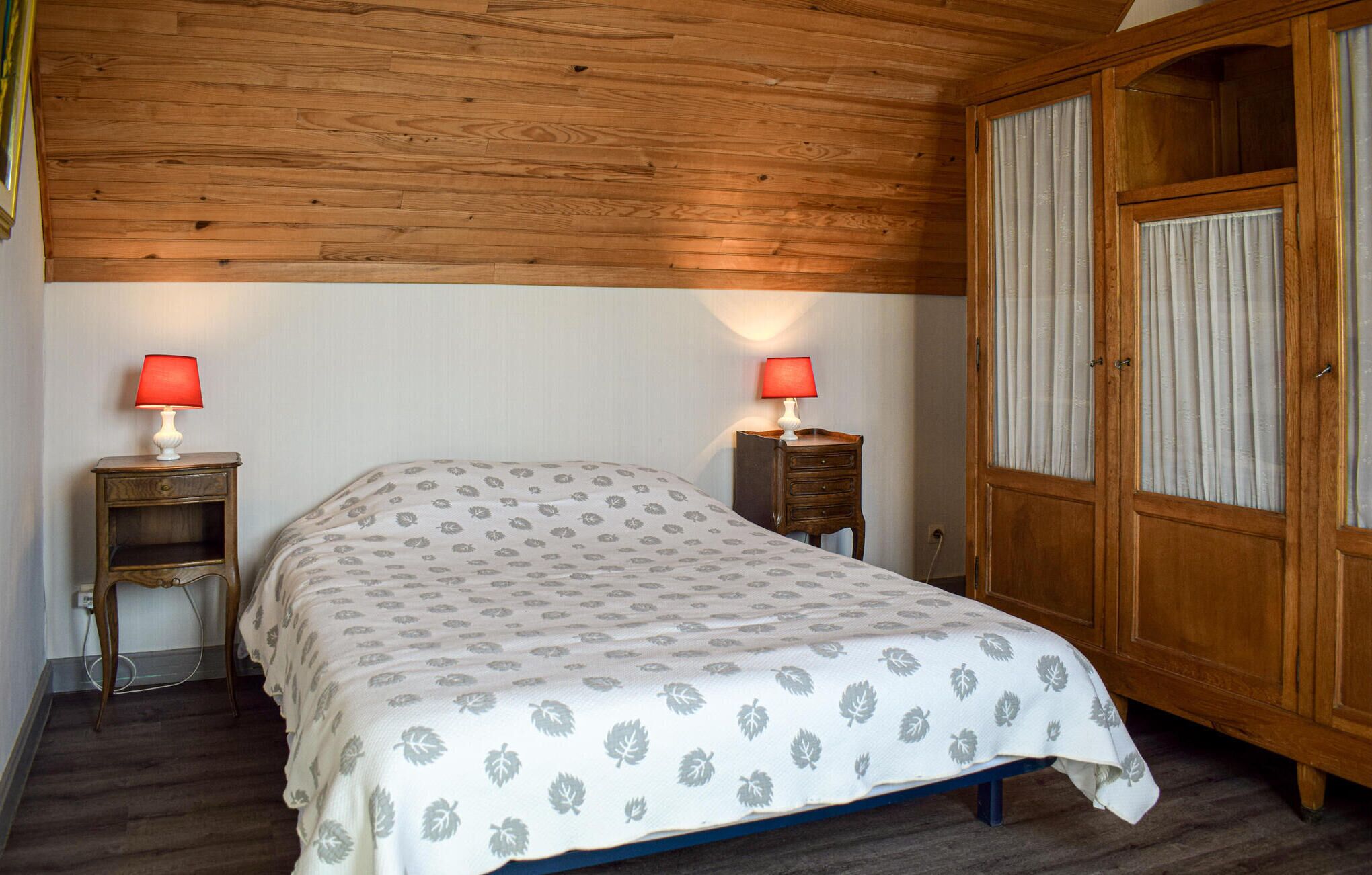 Een slaapkamer met een houten plafond, een bed met een bedsprei met dessin, twee nachtkastjes met lampen en een inloopkast met gordijnen.