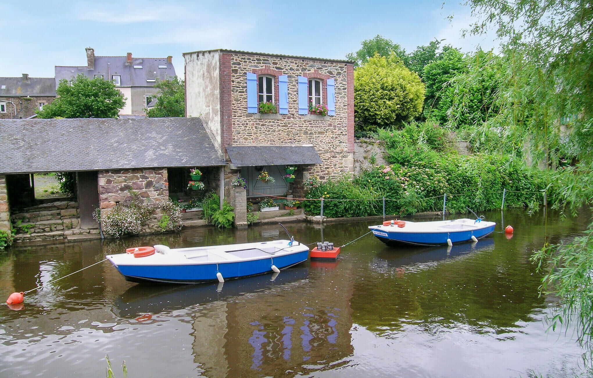 Two boats moored along a canal, with a stone building featuring blue shutters and hanging flower baskets in the background.