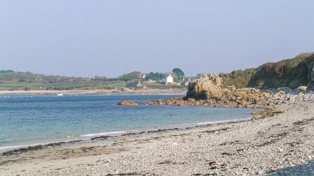 Une plage côtière au rivage de galets, avec un promontoire rocheux et des maisons en arrière-plan.