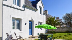 Une maison blanche avec une porte et des fenêtres bleues, un parapluie vert et du mobilier d’extérieur.