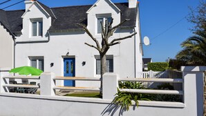 Une maison blanche à deux étages, avec une porte bleue, une antenne parabolique et un arbre dans le jardin.