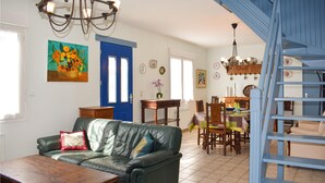 Un vaste salon doté d’un canapé en cuir vert, d’une table basse en bois et d’un coin repas avec une table et des chaises en bois. Un escalier bleu est visible à l’arrière-plan.