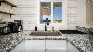 A kitchen with white subway tiles, a granite countertop, a double sink, a coffee maker, and a window with a view of a desert landscape. - Wellness Retreat w ColdPlunge, HotTub & Redlight Sauna (Twentynine Palms)