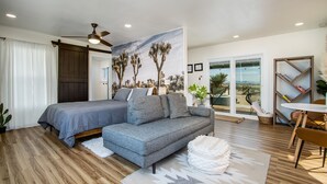 A bedroom with a bed, a gray sofa, a ceiling fan, and a mural of desert trees. - Wellness Retreat w ColdPlunge, HotTub & Redlight Sauna (Twentynine Palms)