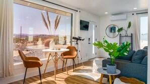 A modern living room with a large window, a telescope, and a potted plant. - Wellness Retreat w ColdPlunge, HotTub & Redlight Sauna (Twentynine Palms)
