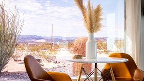 A room with a large window, a white vase with pampas grass, two brown chairs, and a small round table. - Wellness Retreat w ColdPlunge, HotTub & Redlight Sauna (Twentynine Palms)