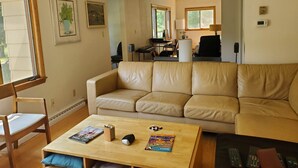 Una sala de estar con un sofá de cuero color beige, una mesa de centro de madera con una revista, un control remoto y una lámpara. Se observa un escritorio con una computadora y una silla al fondo.