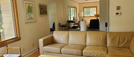 En stue med en beige lædersofa, et træbord med et magasin, en fjernbetjening og en lampe. I baggrunden er der et skrivebord med en computer og en stol.