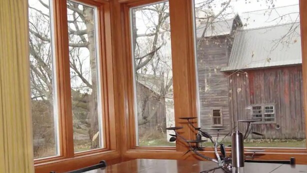 Un comedor con una gran mesa de madera y sillas, ventanas amplias con vistas a una construcción de madera rústica y árboles.