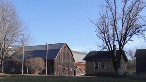 Un entorno rural con un gran granero de madera, una casa más pequeña y un árbol en primer plano.