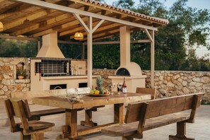 An outdoor patio with a wooden table and benches, a stone wall, and a barbecue grill.