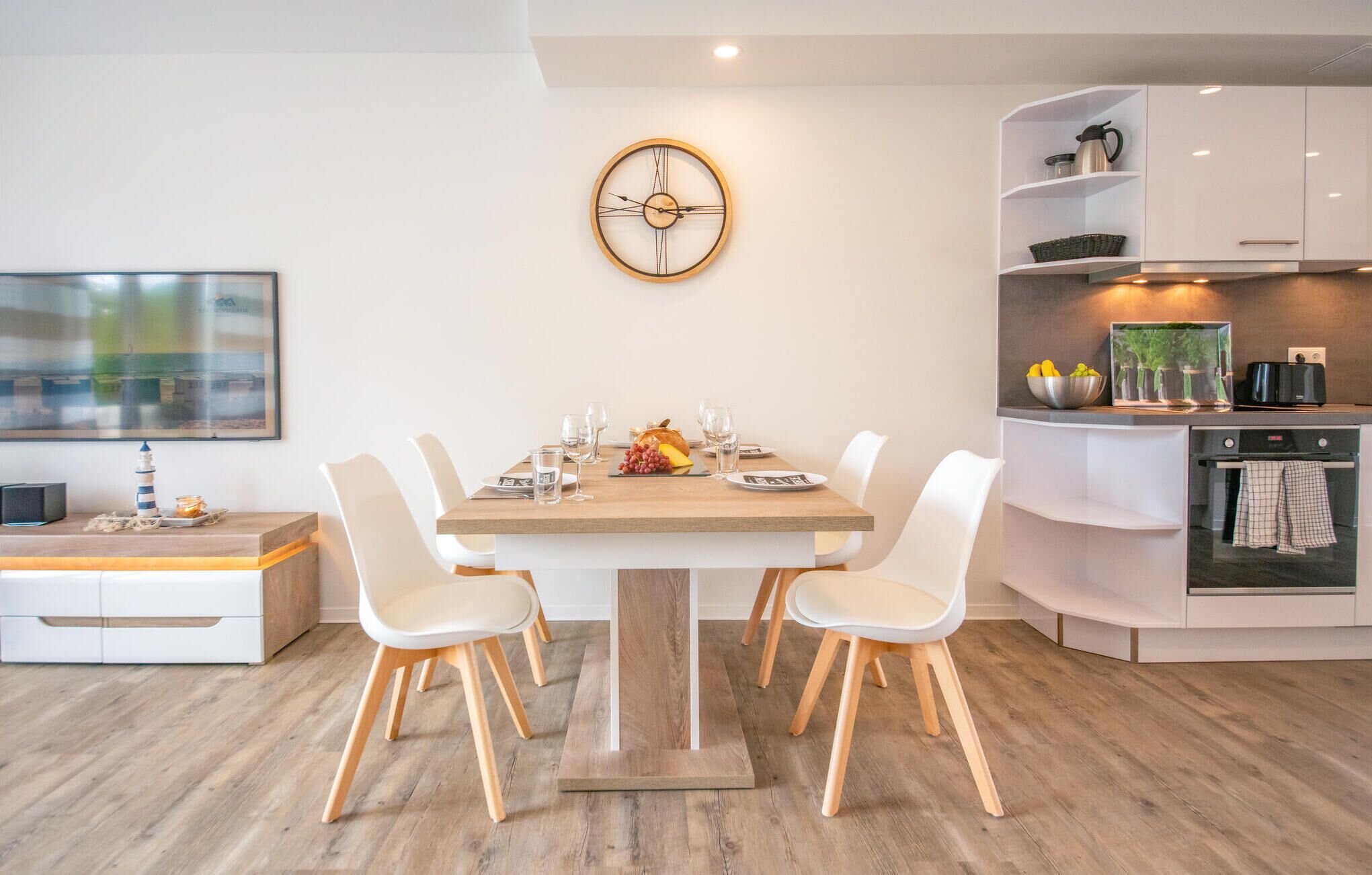 A modern kitchen with white cabinetry, a wooden dining table with four chairs, and a wall-mounted clock.