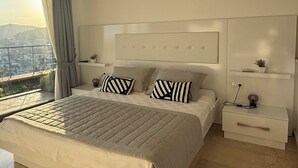 Un dormitorio moderno con una cama grande, mesitas de noche, un buró y aire acondicionado instalado en la pared, con vistas a la ciudad a través de un ventanal amplio.