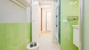 Un couloir étroit aux murs recouverts de carreaux verts et blancs, un radiateur blanc, une petite table blanche avec des serviettes, et une porte au fond.