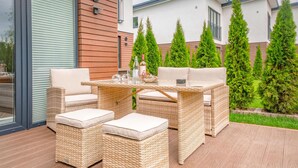 Un coin salon extérieur aménagé avec des meubles en osier, des coussins, une table à plateau en verre et une bouteille de vin.