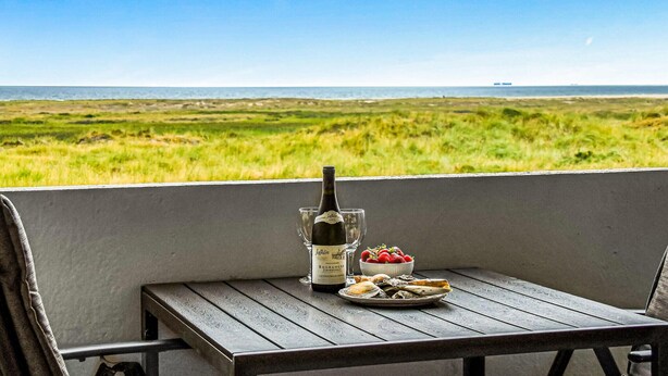 Una mesa con una botella de vino, un bol de fresas y un plato de comida en un balcón con vistas a un campo y al mar.