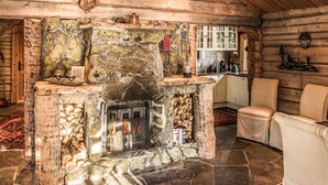 A stone fireplace with a log fire, wooden walls, a kitchen area, and a seating area with chairs.