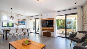 Uma sala de estar moderna com uma mesa de centro de madeira, uma TV fixada em uma parede de madeira, e uma varanda com um sofá e uma mesa.