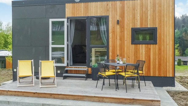 A modern, compact building with a wooden exterior and large glass windows. It features a small outdoor seating area with two yellow chairs and a table, a sliding glass door entrance, and a small window with a view of greenery.