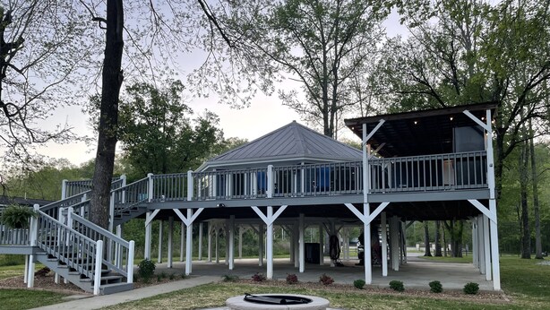 A covered outdoor deck with a stone fire pit in the center, surrounded by a grassy area and trees. - The Tree House! (Florence)