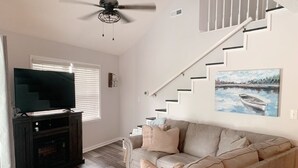 A living room with a beige sofa, a television on a dark wooden stand, a ceiling fan, a staircase, and a painting of a boat on water. - The Tree House! (Florence)
