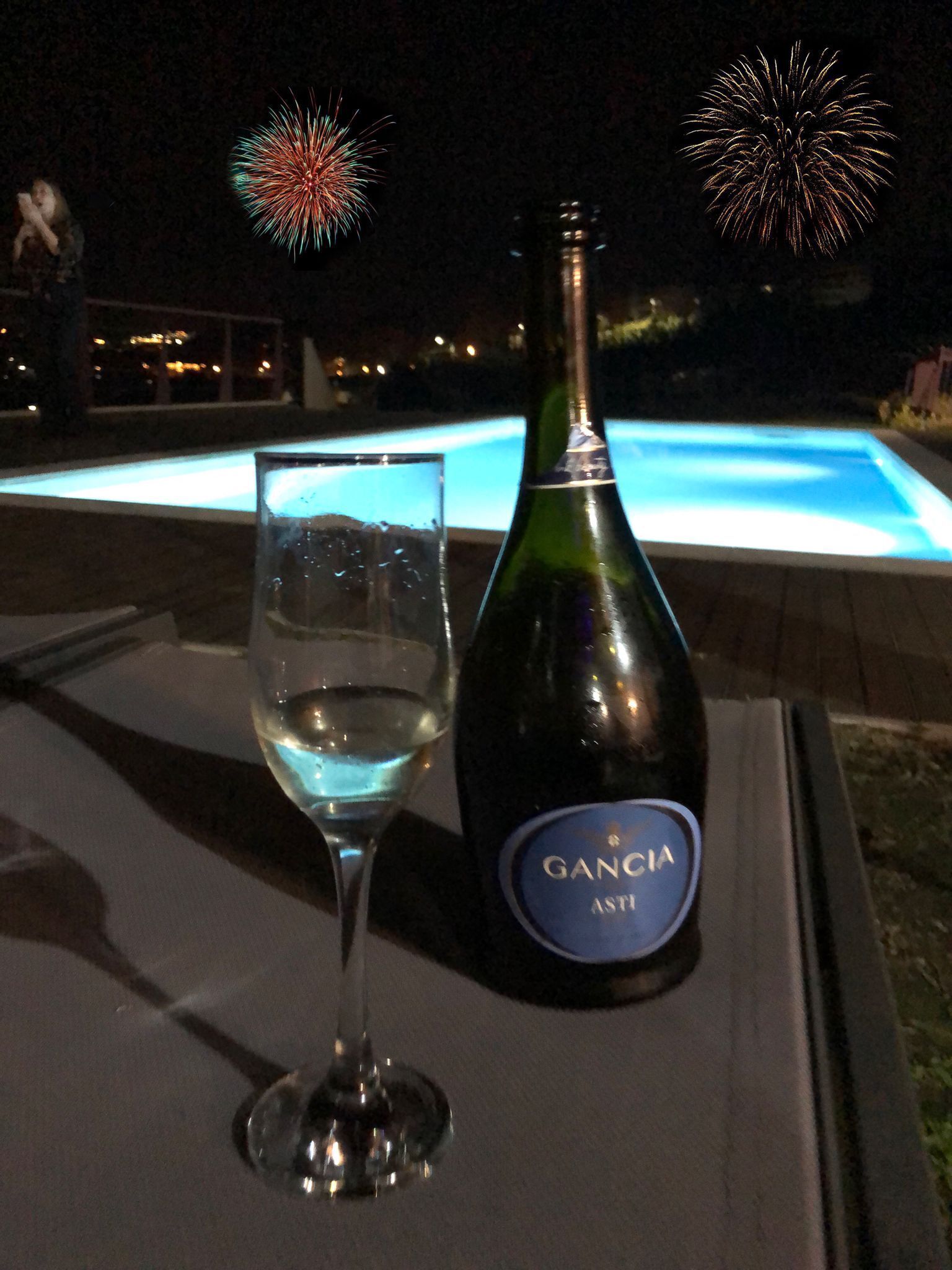 En flaska Gancia Asti-vin och ett glas på ett bord, med en pool och fyrverkerier i bakgrunden.