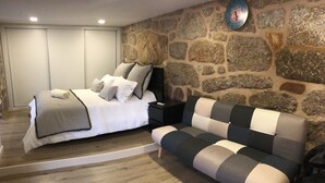 Quarto com cama, mesa de cabeceira, sofá, parede de pedra e luminária no teto.