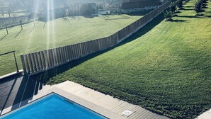 Piscina com deck de madeira e vista para um campo gramado e um edifício ao fundo.