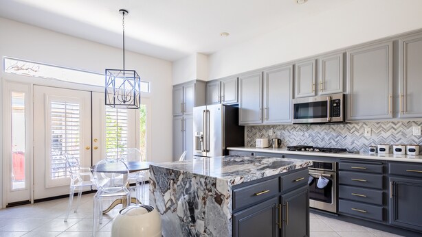 A modern kitchen with grey cabinets, a marble island, stainless steel appliances, and a herringbone tile backsplash. - Luxury, spacious, spa home, sleeps 8, Chef Kitchen, office, and separate casita. (Palm Desert)