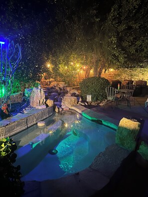 Area piscina esterna illuminata di notte, con rocce, una piccola cascata e posti a sedere.