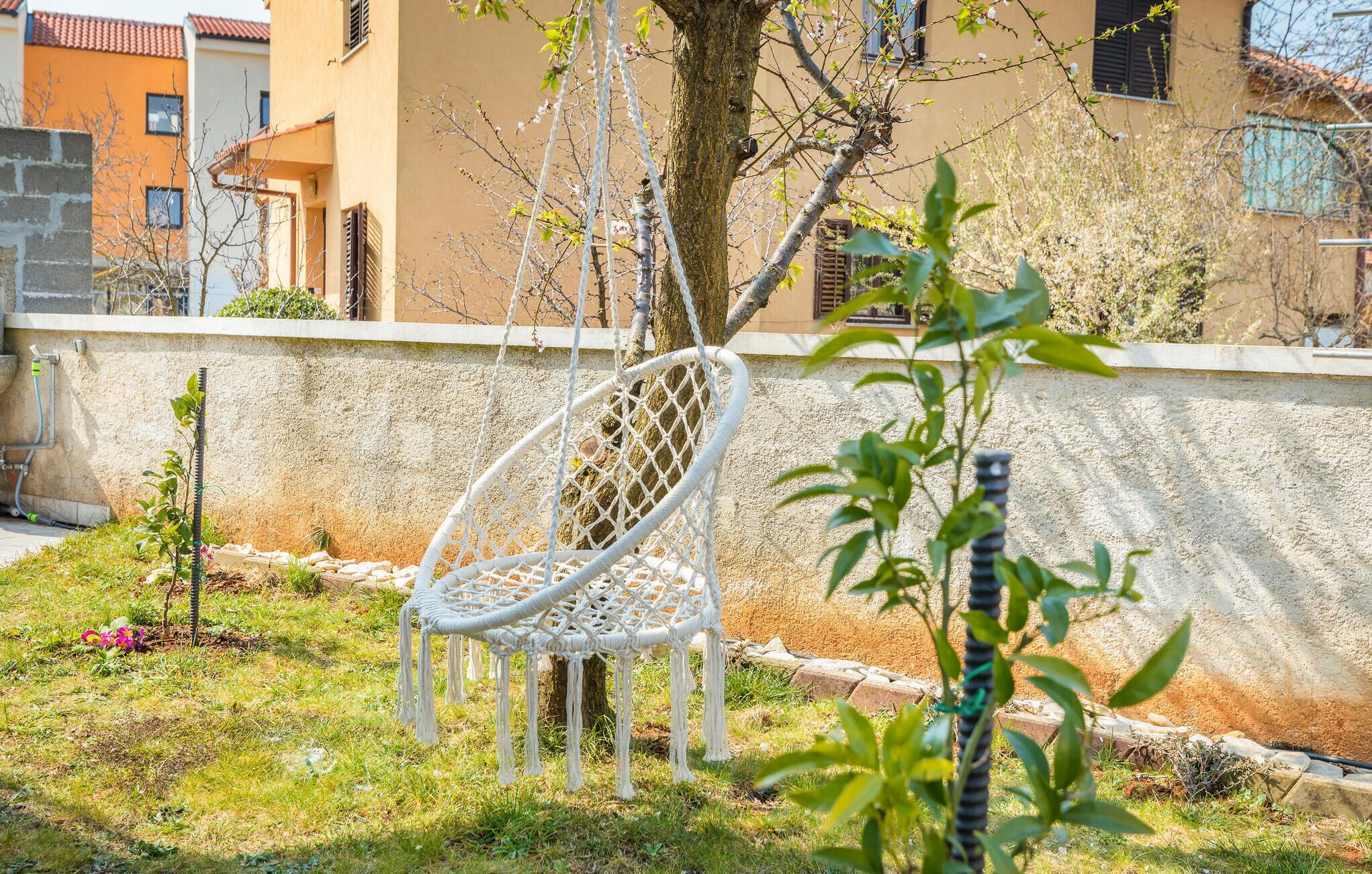 A white macrame chair in a garden with a tree and a small plant.