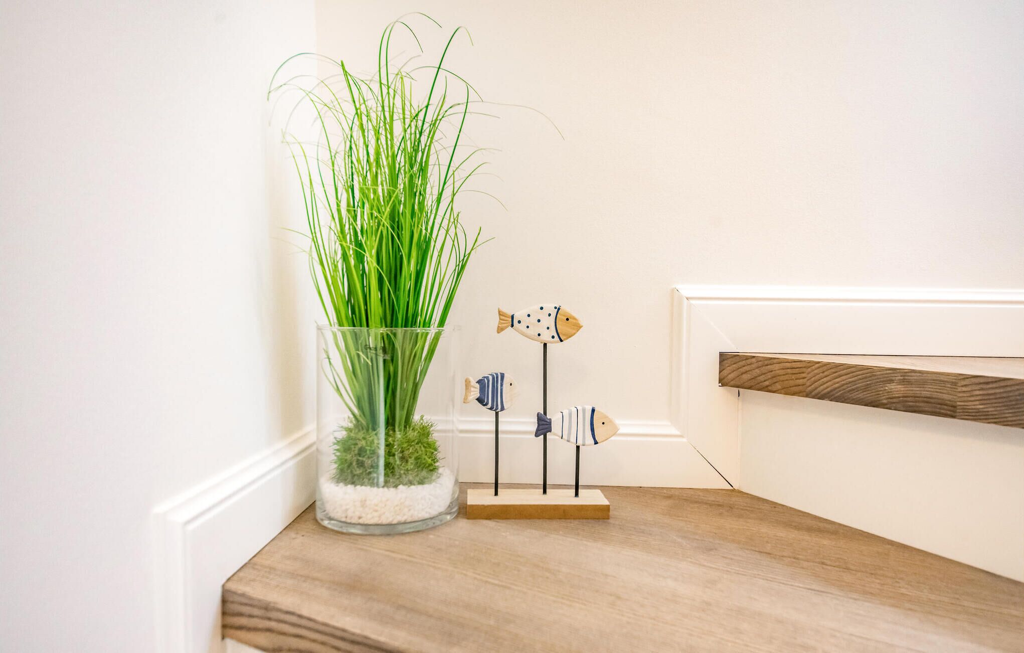 A clear glass vase with green grass and a small wooden stand holding three fish-shaped decorations.