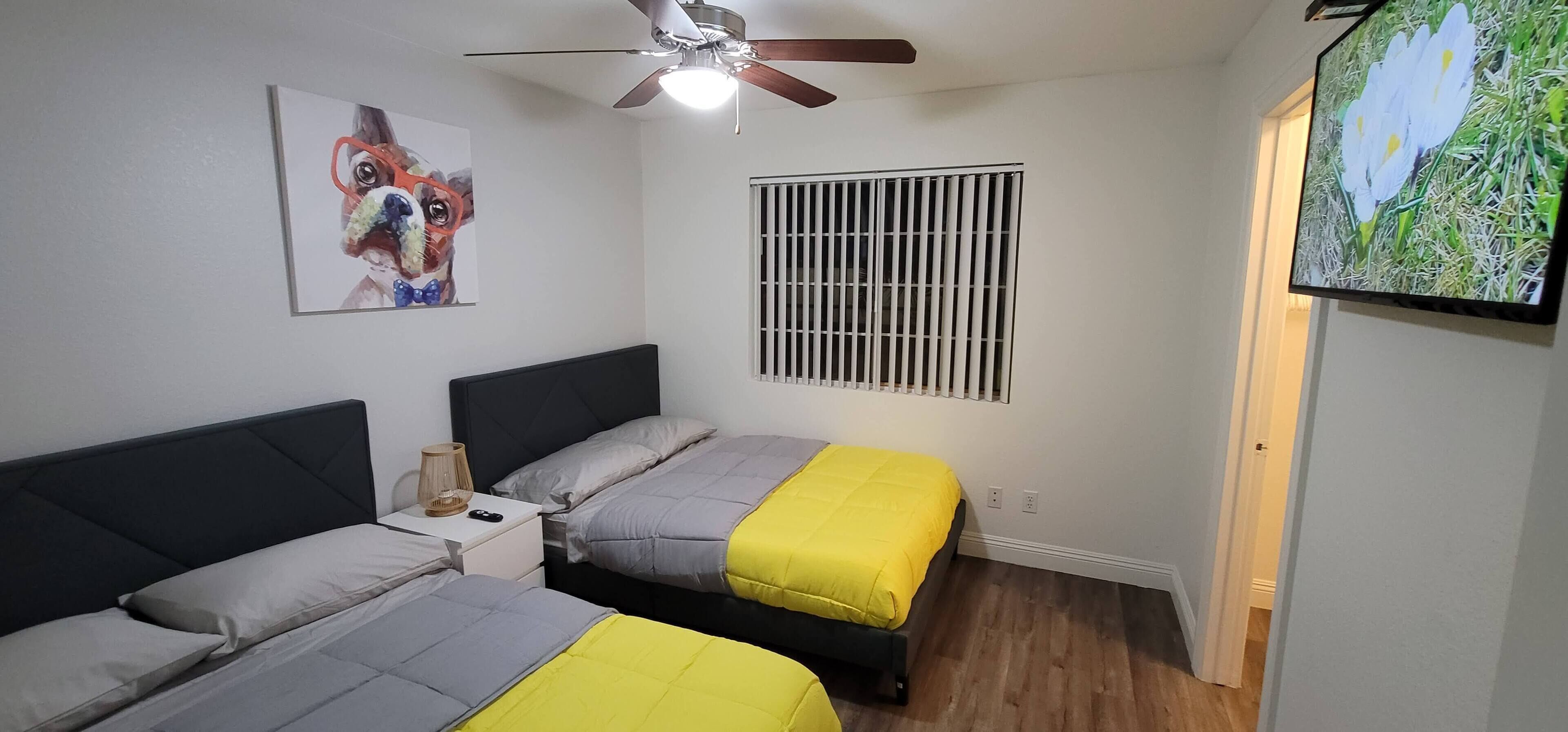 A bedroom with two beds, a ceiling fan, a window with bars, and a TV on the wall.