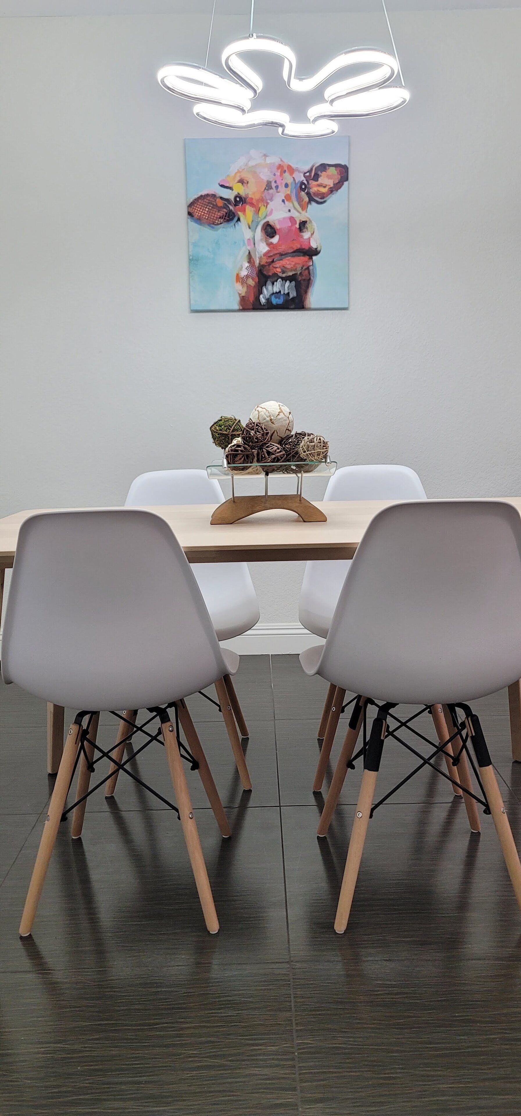 A dining area with a wooden table, two white chairs, a modern pendant light, and a colorful painting on the wall.
