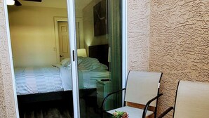 Ein kleiner Balkon mit einer Schiebetür, die zu einem Schlafzimmer führt. Dort steht ein Tisch mit einer Blumenvase und ein Stuhl.