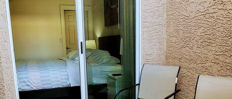 Un pequeño balcón con una puerta corredera de vidrio que da a un dormitorio. Hay una mesita con un florero y una silla.