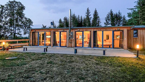 Een moderne houten chalet met grote glazen deuren, een verhard terras en een bankje.