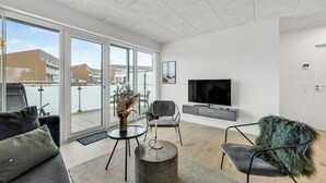 En moderne stue med stort fjernsyn, behagelige siddepladser, en balkon med udsigt til bygninger og et stort vindue.