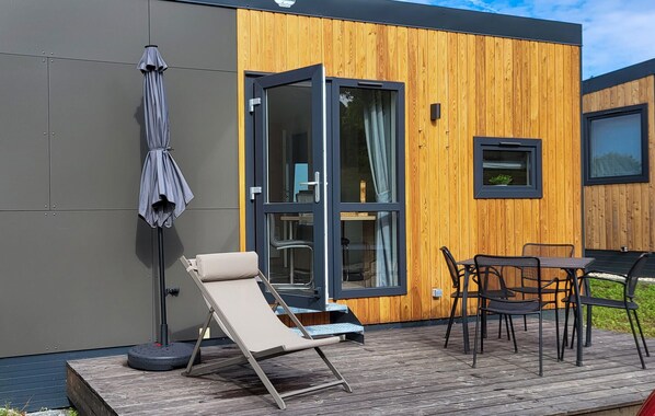 A modern outdoor seating area with a sun lounger, a table, and chairs, adjacent to a wooden building with a glass door.
