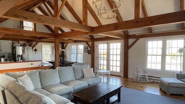 A spacious living room with a large sectional sofa, a coffee table, and a dining area with a table and chairs. The room features exposed wooden beams and large windows allowing natural light. - 4 Bedroom, Upside Down Post and Beam Nantucket Home (Nantucket)
