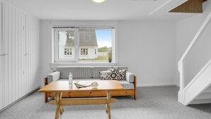 A room with a wooden table, a sofa with patterned cushions, a window with a view of a house, and a staircase. - Lovely home in Thisted with kitchen (Thisted)