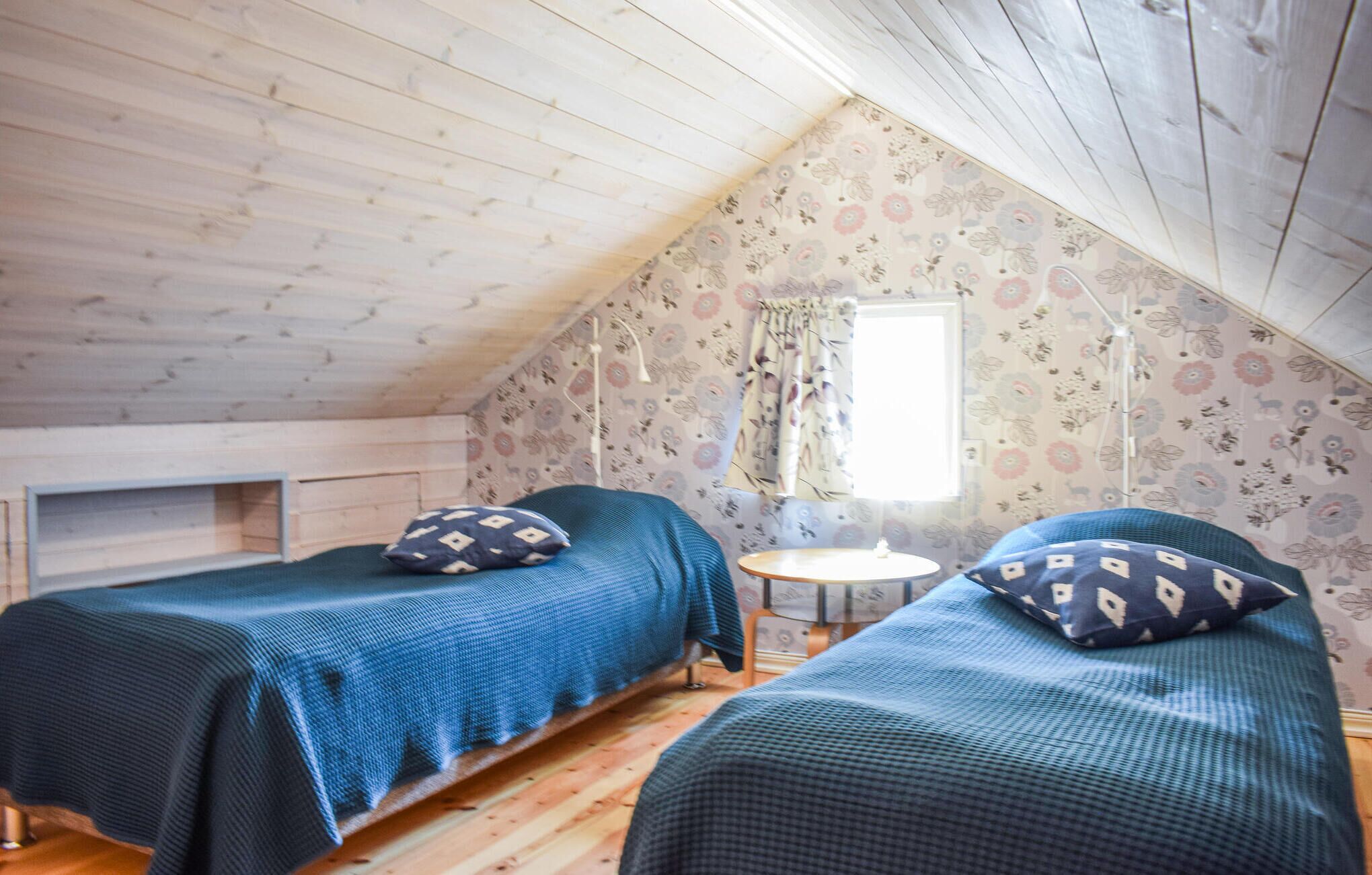 Ein kleines, gemütliches Schlafzimmer im Dachgeschoss mit zwei Einzelbetten, einem kleinen Tisch und einem Fenster mit blumigen Vorhängen.