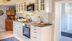 Eine kleine Küche mit weißen Schränken, einem eingebauten Backofen und einer Mikrowelle. Ein Holzregal zeigt verschiedene Gegenstände, die Wände sind mit ornamentalem Wandpapier versehen.